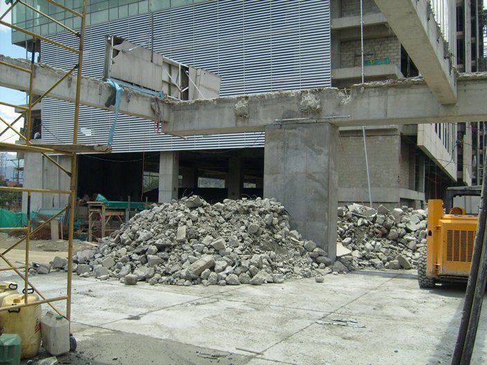 Montón de escombros frente a una columna de hormigón. Edificio en construcción/demolición; vehículo de construcción amarillo visible.