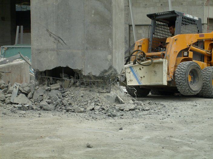 Minicargadora amarilla demoliendo un pilar de hormigón, escombros en el suelo.
