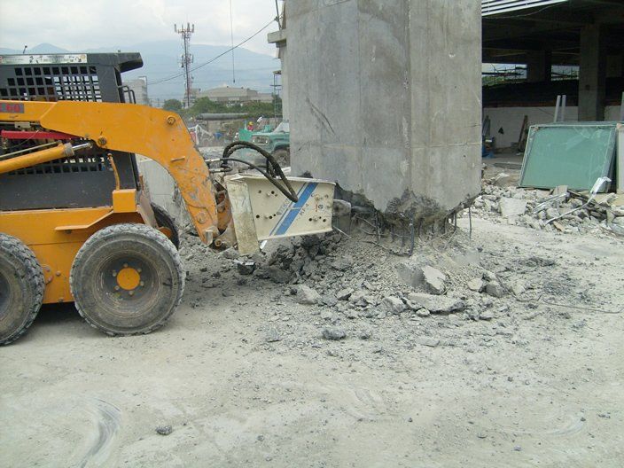 Minicargadora amarilla con una herramienta de demolición rompiendo un pilar de hormigón en un entorno de construcción al aire libre.