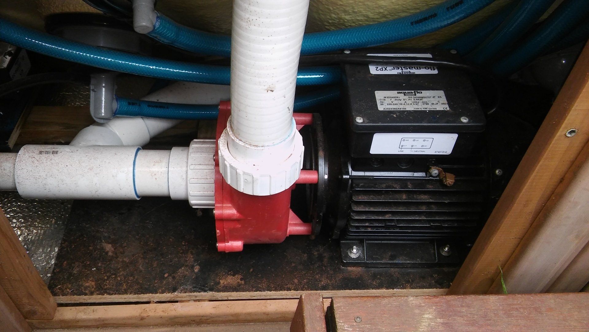 Under-sink plumbing with white PVC pipes, a red valve, and a black pump unit in a wooden cabinet