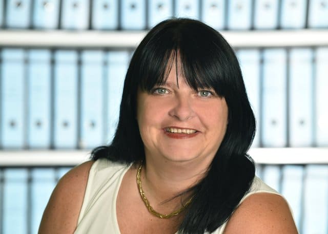 Eine Frau mit langen schwarzen Haaren lächelt vor einer Reihe von Ordnern.
