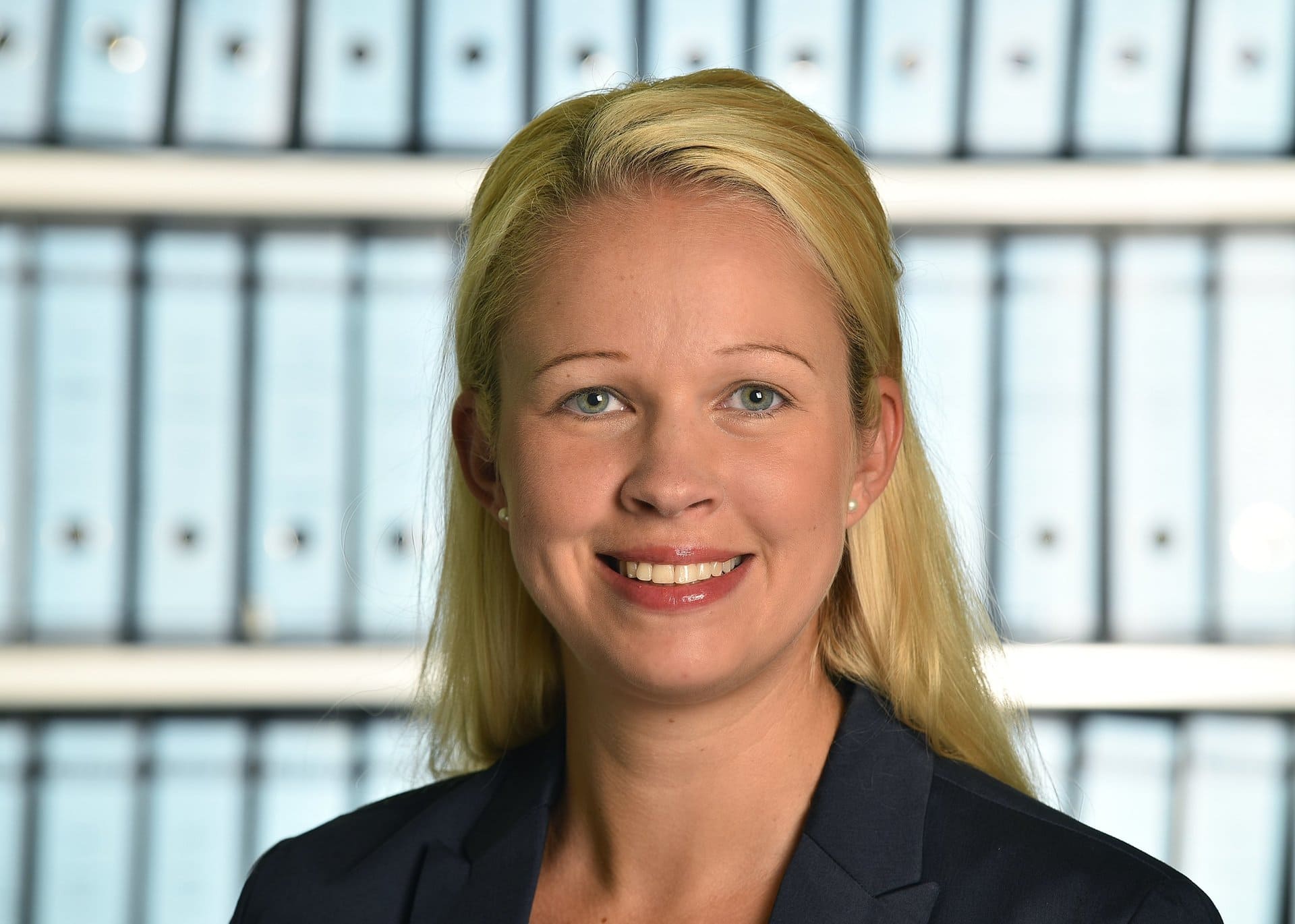 Eine Frau im Anzug lächelt vor einer Reihe von Ordnern.