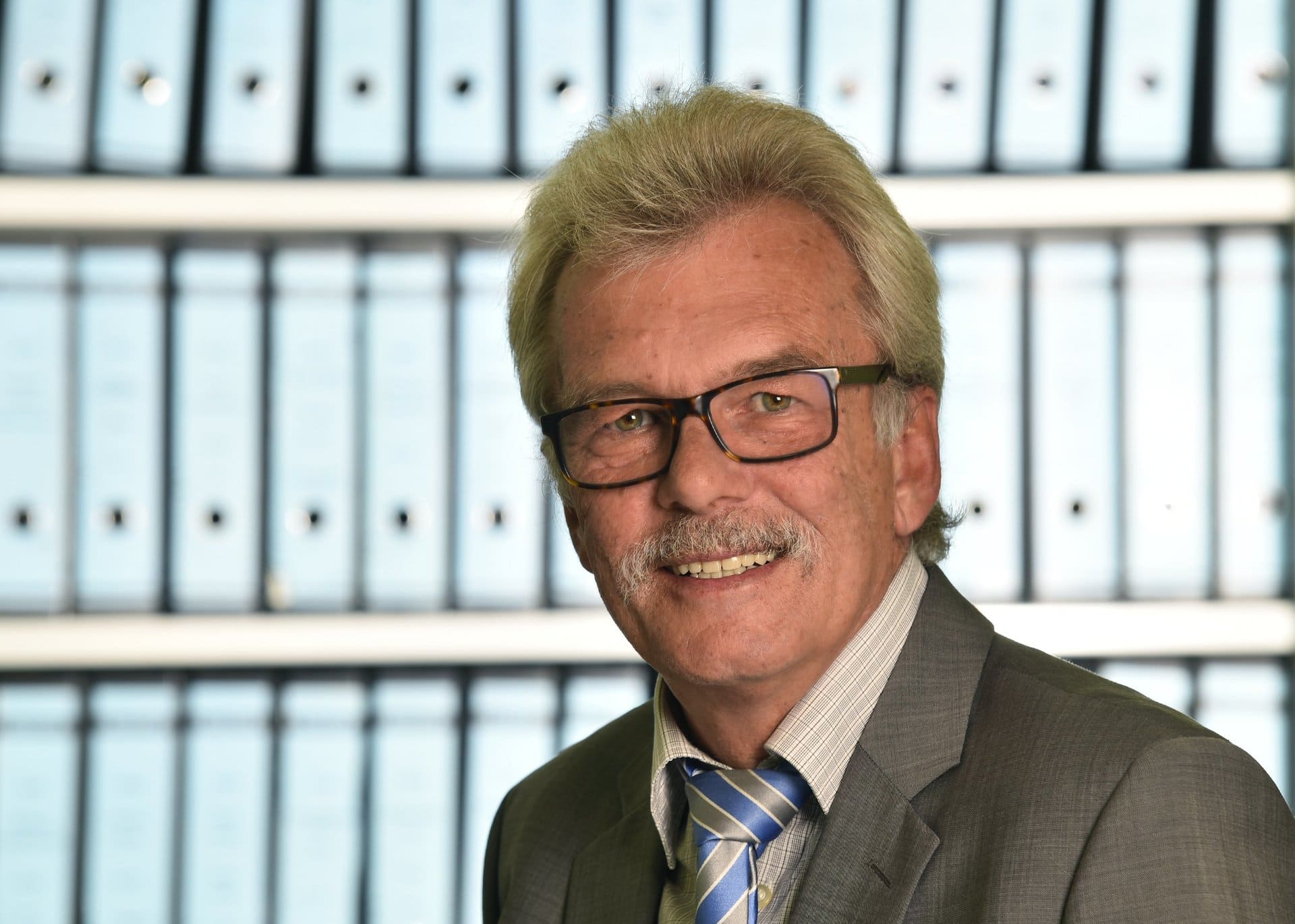 Ein Mann in Anzug und Krawatte lächelt vor einer Reihe von Ordnern.