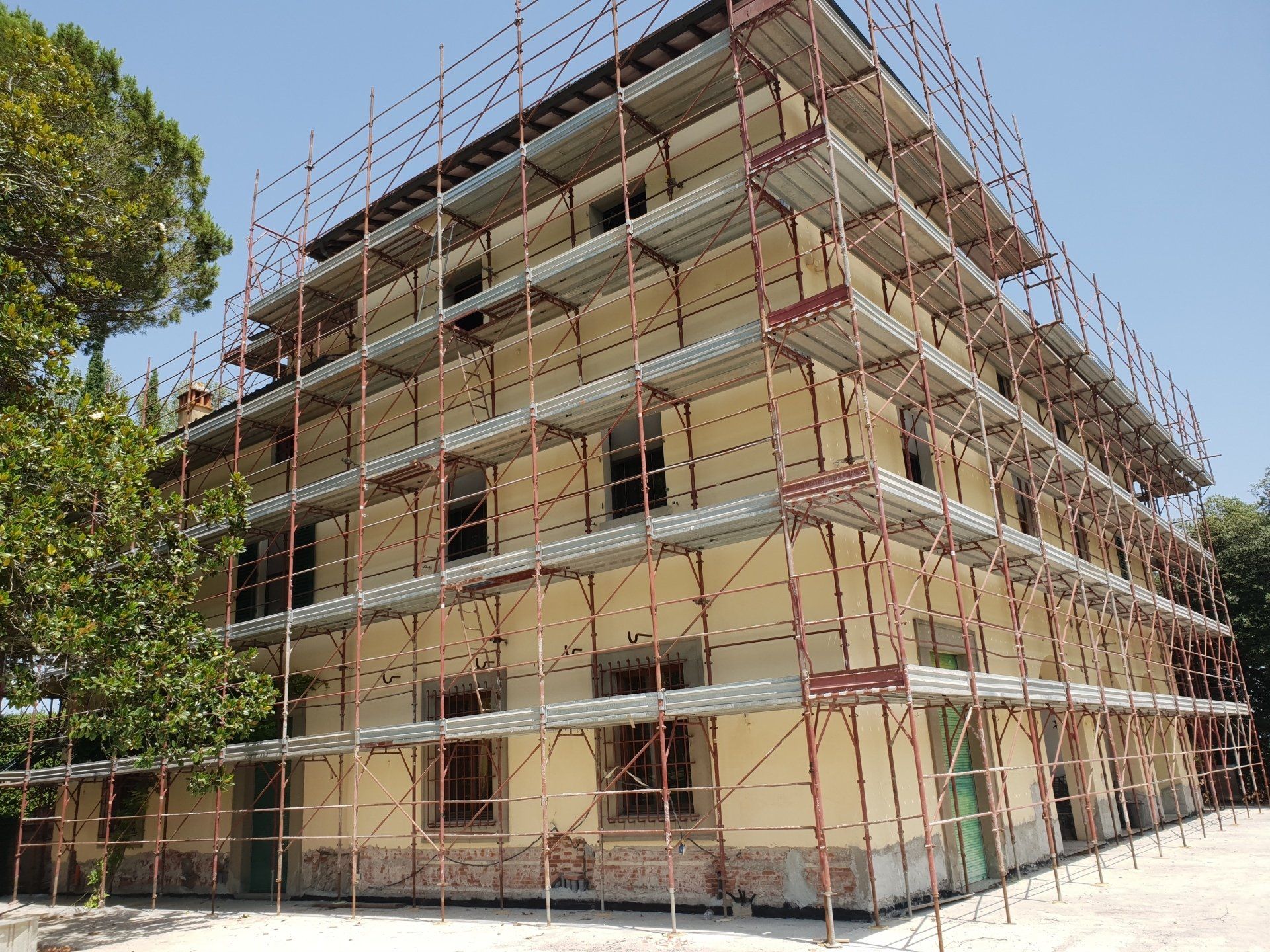 Edificio in fase di ristrutturazione, coperto di impalcature; pareti beige, infissi marroni, giornata di sole.