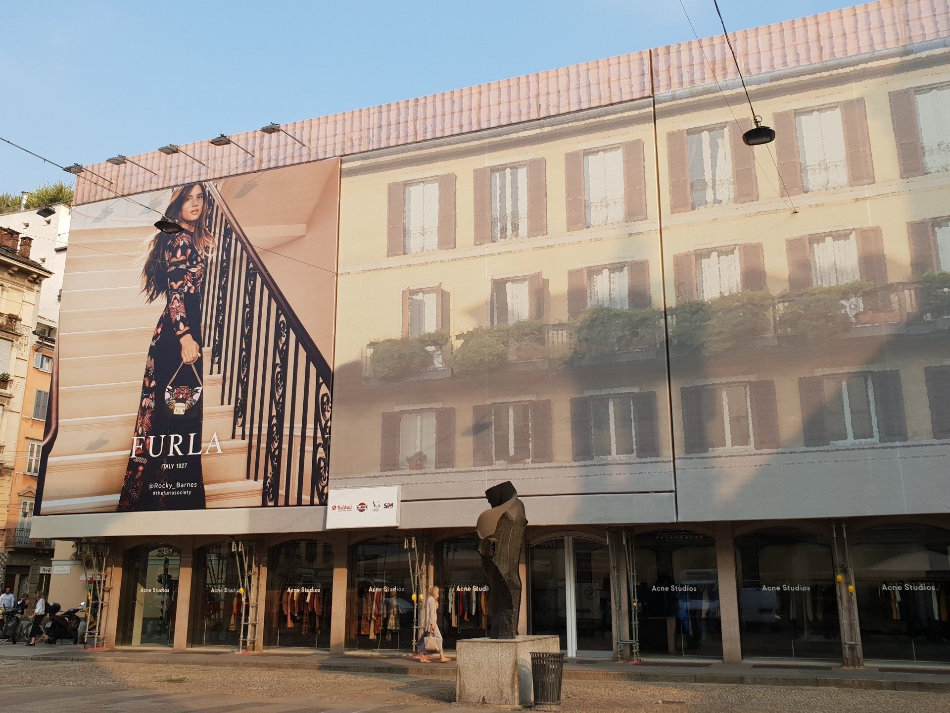 Edificio con grande striscione pubblicitario, negozi sottostanti. Donna davanti alla statua.