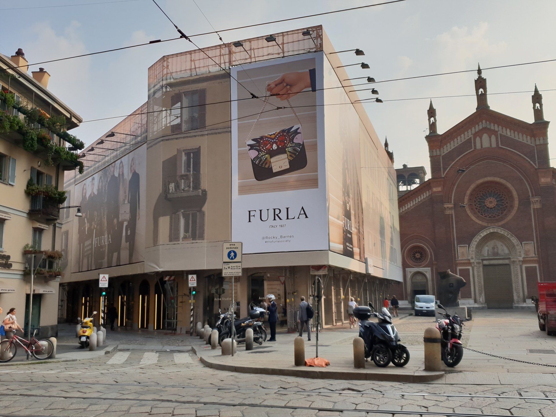 Esterno di un negozio Furla a Milano, con una grande pubblicità e una chiesa sullo sfondo. Motociclette e persone in strada.