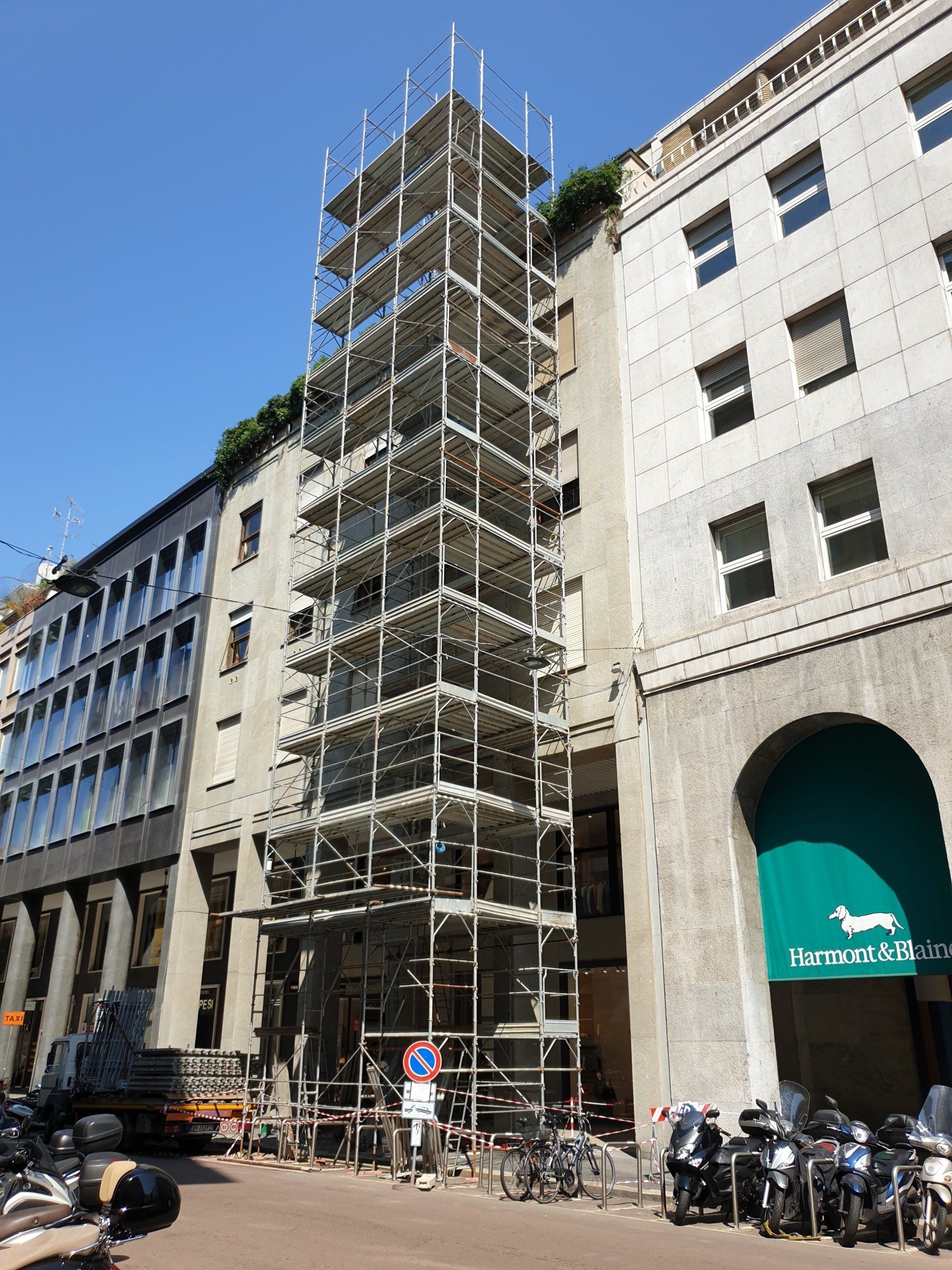 Impalcature sulla facciata di un edificio accanto a una banca con tenda da sole verde; motociclette parcheggiate. Giornata di sole.