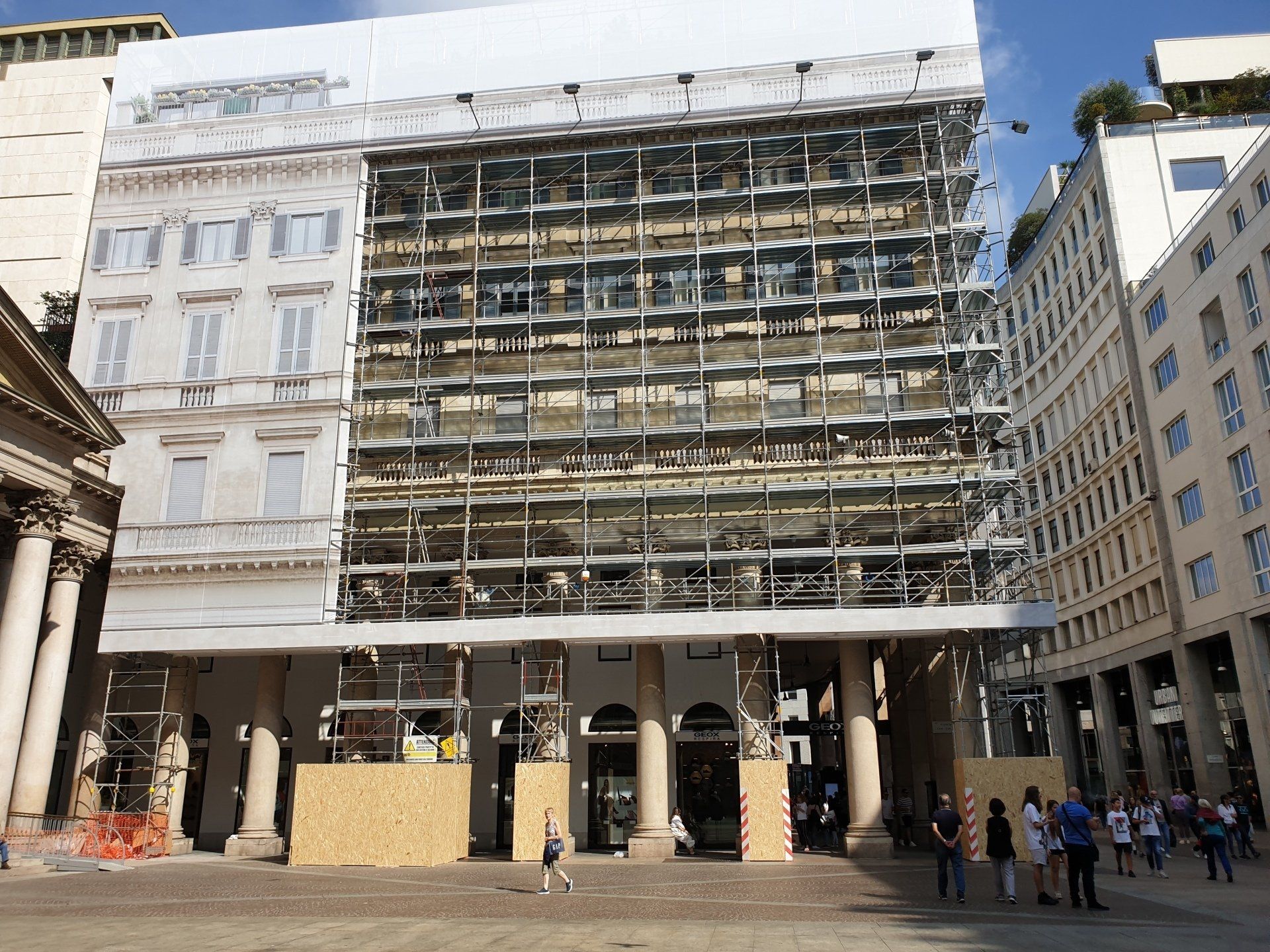 Edificio in ristrutturazione, coperto da impalcature, in una piazza cittadina. Persone che camminano nelle vicinanze, giornata di sole.