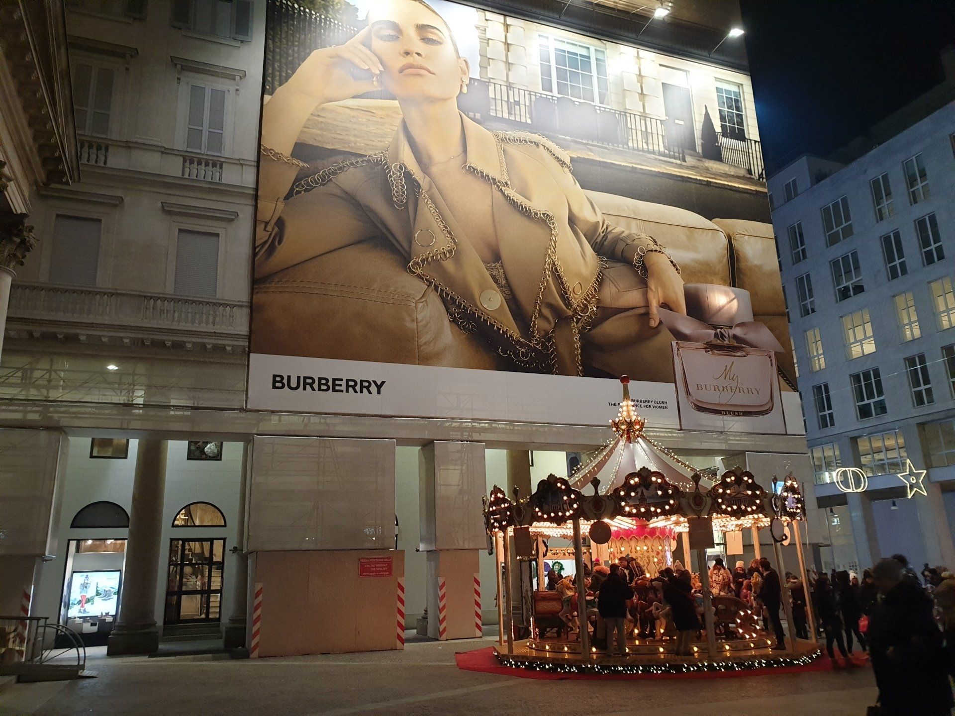 Un grande cartellone pubblicitario di Burberry su una giostra. L'annuncio mostra una donna con una giacca marrone chiaro. Ambientazione serale.