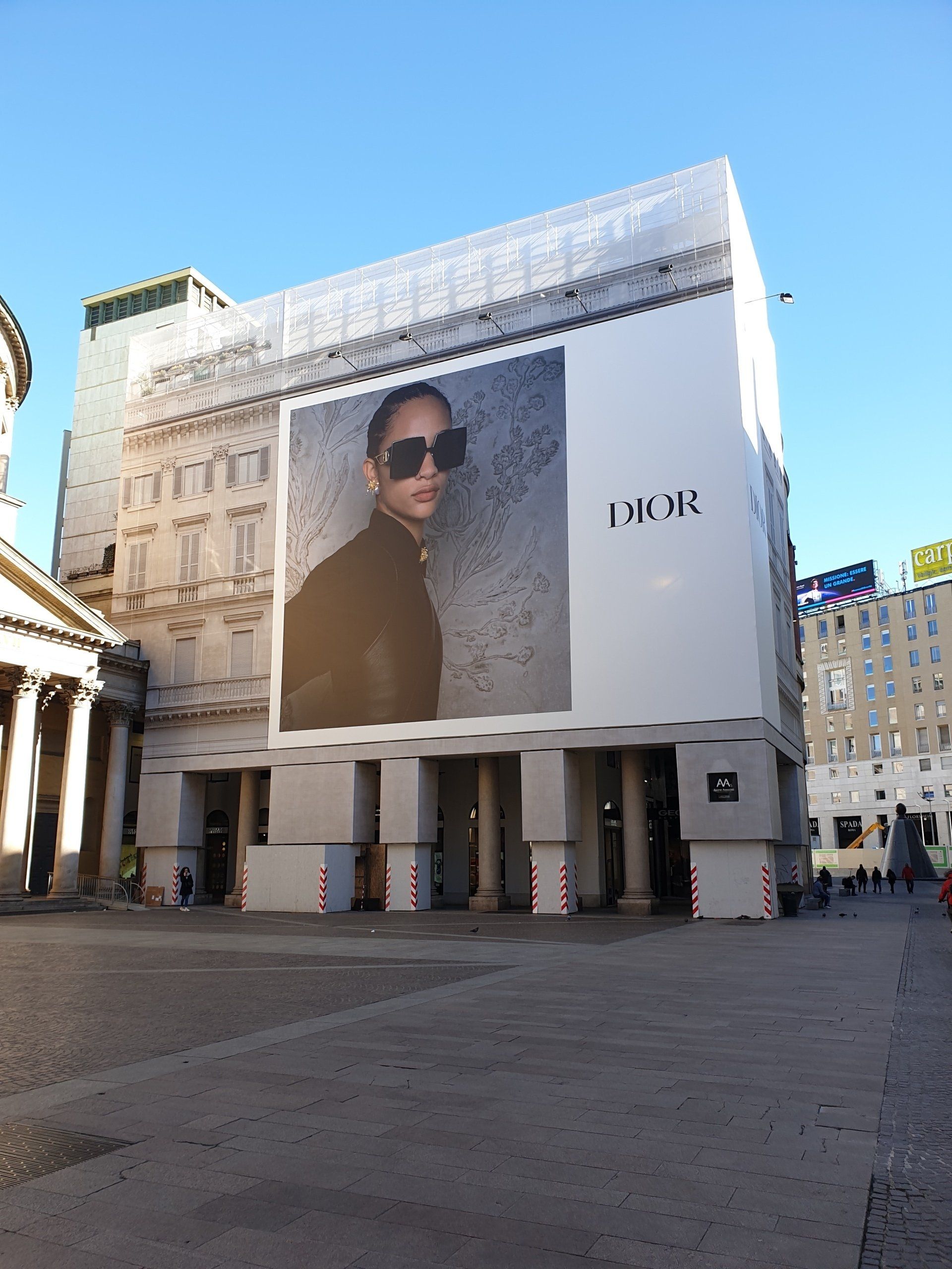 Grande pubblicità Dior sulla facciata di un edificio in una piazza pubblica. Persona con occhiali da sole, colonne architettoniche visibili.