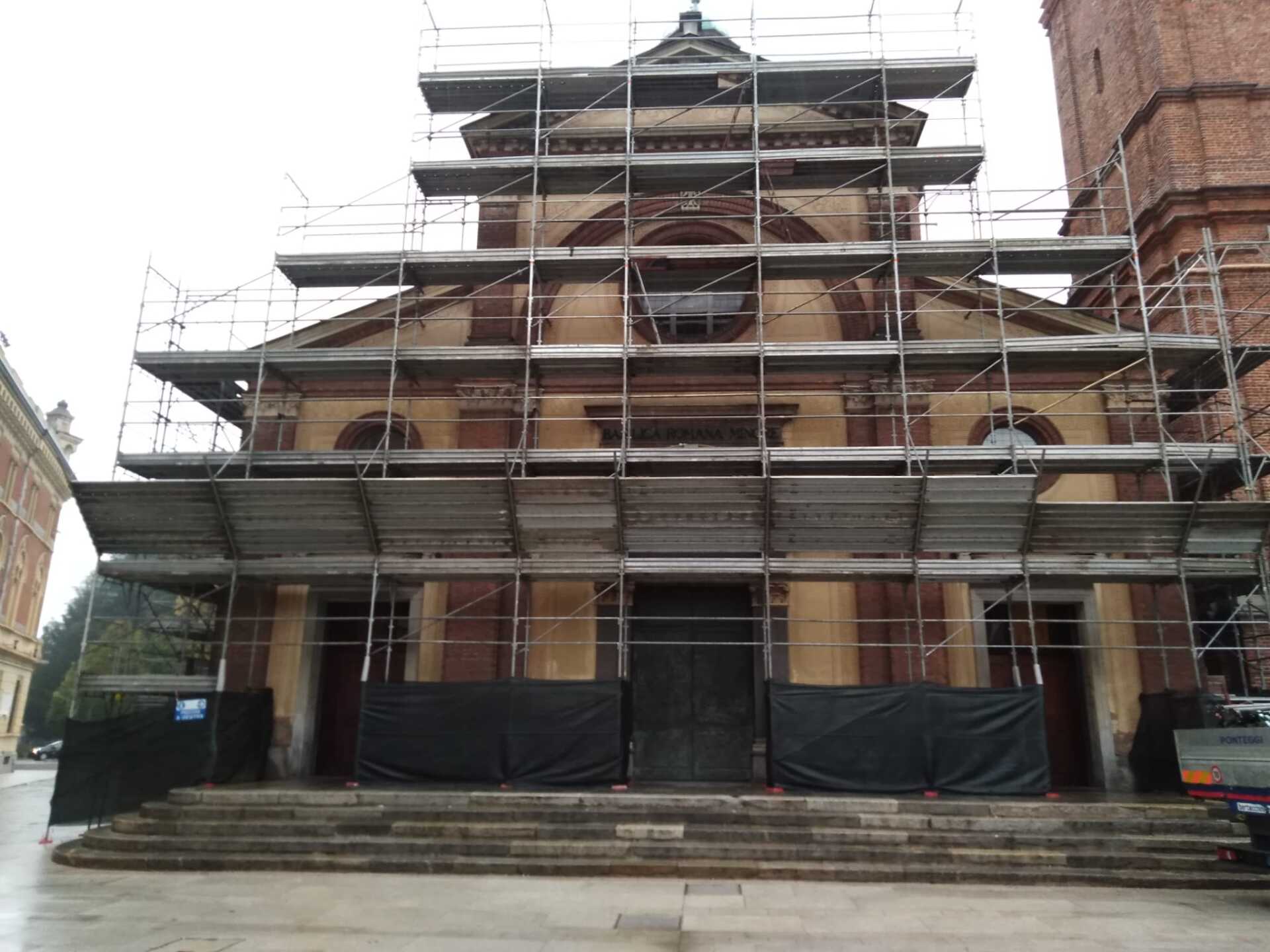 Facciata di una chiesa in costruzione con impalcature. Edificio marrone e beige con porte e gradini scuri.