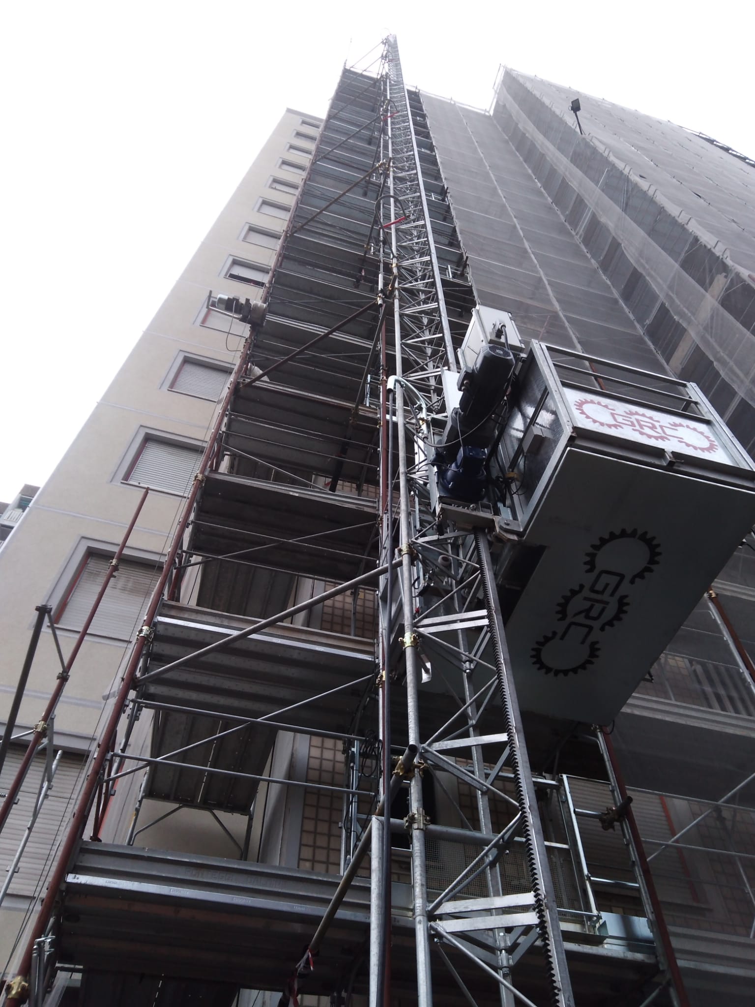 Un ascensore da cantiere collegato a un edificio alto. L'ascensore è bianco e argento.