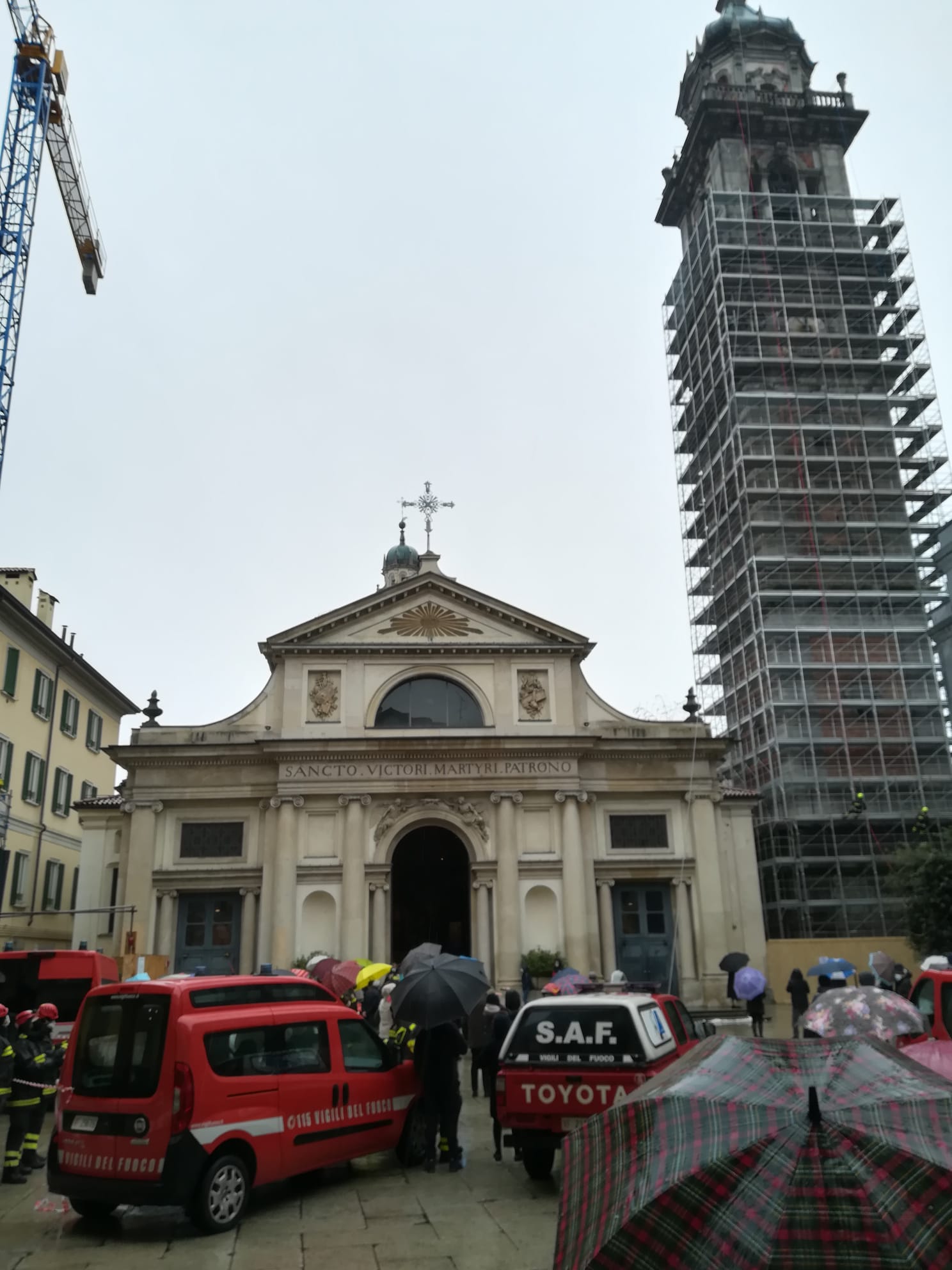 Vista esterna di una chiesa con persone e camion dei pompieri, un'alta torre sotto un'impalcatura e una gru; giornata piovosa.