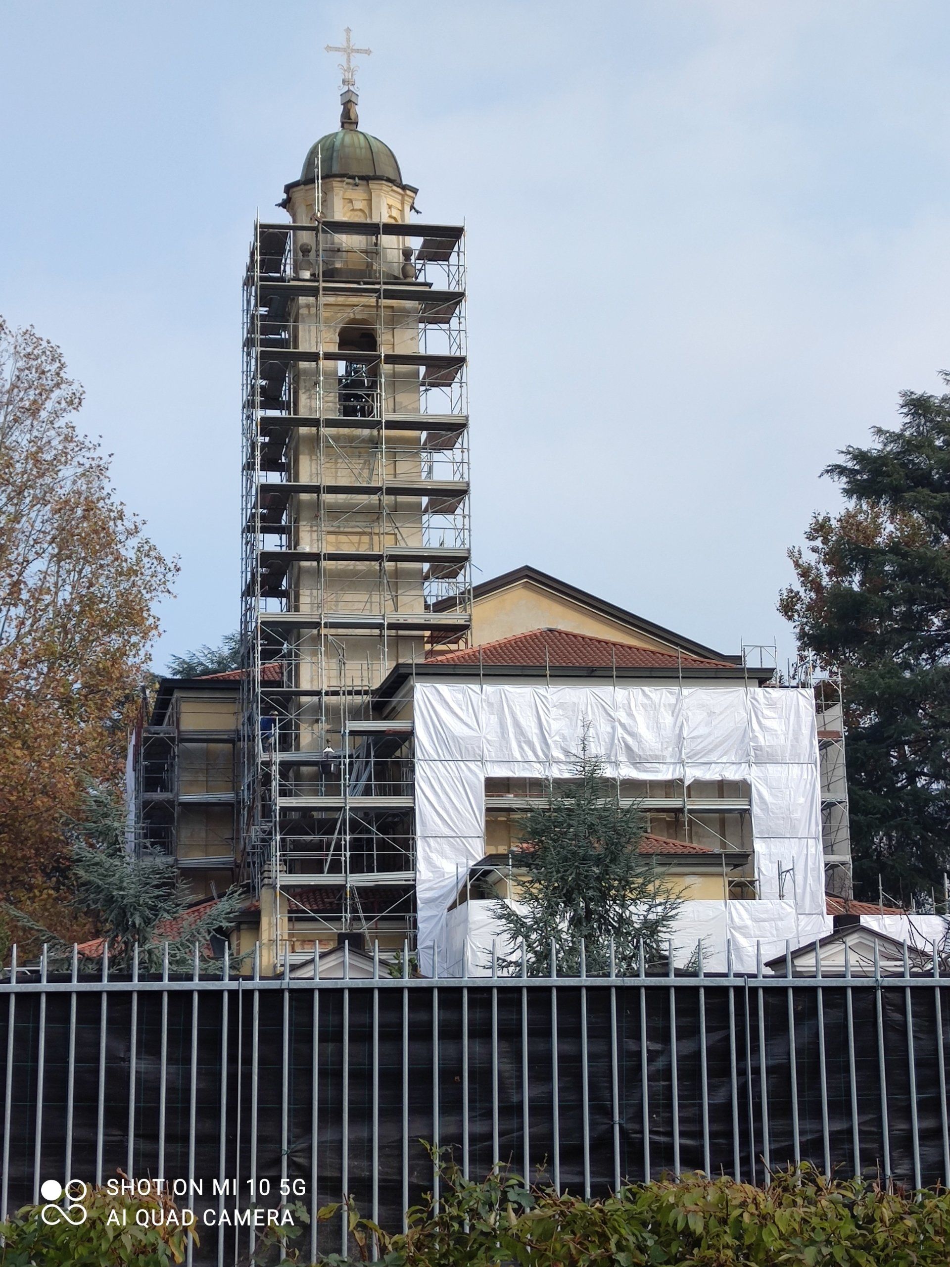 Campanile della chiesa in fase di ristrutturazione, circondato da impalcature e teli protettivi. Cielo grigio.