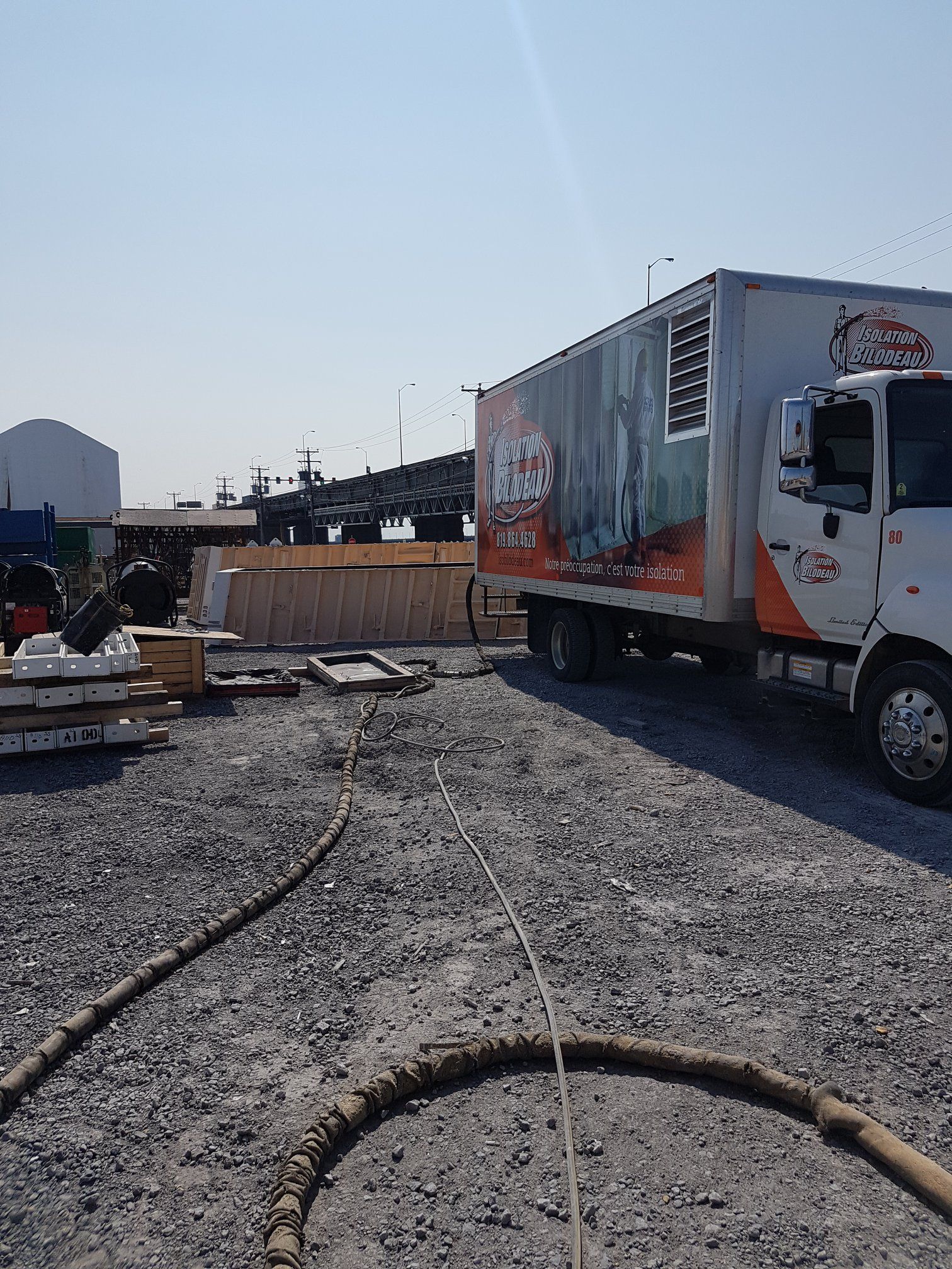 Un camion blanc avec un logo est garé sur du gravier, près de bois et d'équipements. Un tuyau traverse le premier plan.
