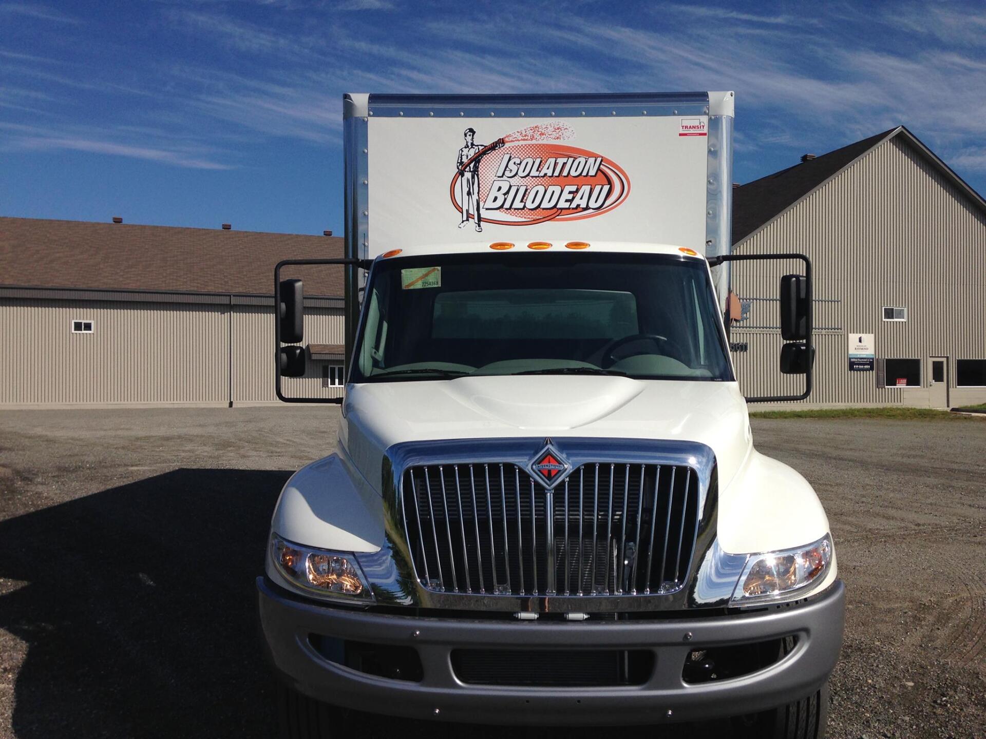 Camion-caisse blanc avec le logo de l'entreprise, garé devant un bâtiment par une journée ensoleillée.