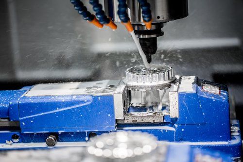 Macchina CNC che fresa un pezzo di metallo argentato, con spruzzatura di refrigerante. Macchina blu con sfondo scuro da officina.