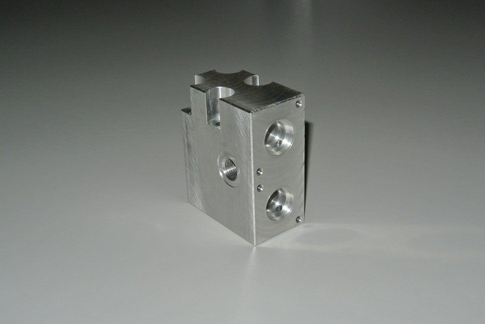 Blocco di metallo argentato con fori e scanalature su una superficie bianca.