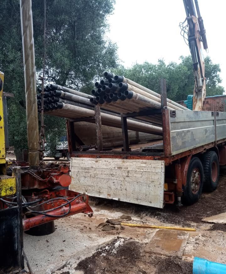 Un camion carico di tubi in un sito di perforazione. Nelle vicinanze c'è una gru e una perforatrice è in funzione.