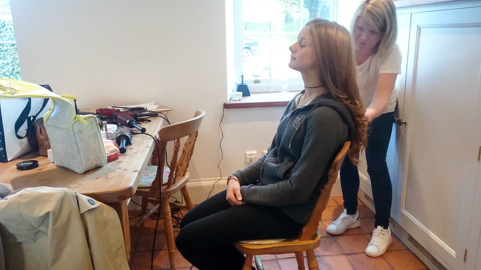 Woman getting hair styled; indoors. Another person stands behind, arranging hair. Table with makeup supplies.