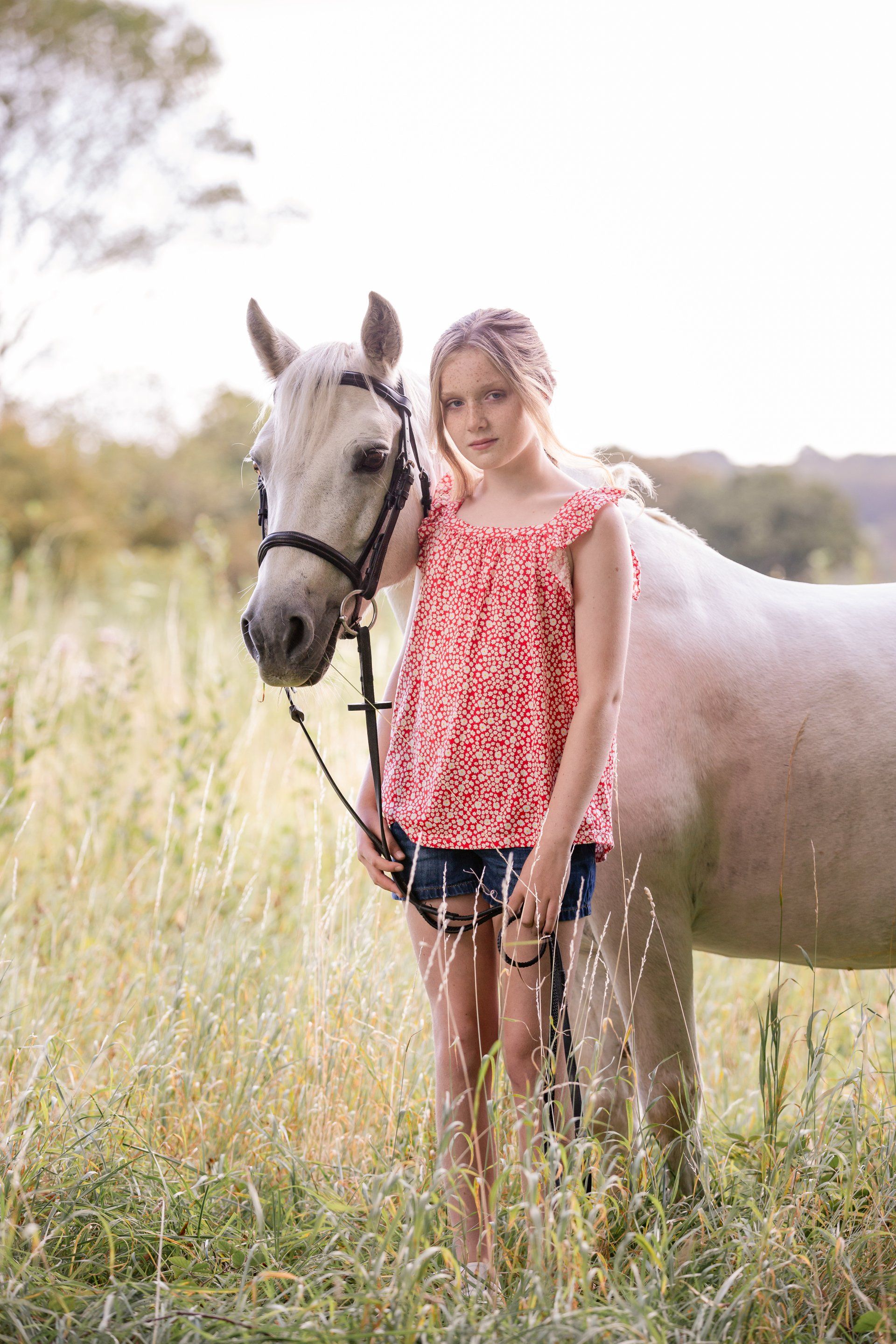 Girl in red top and shorts stands beside a white horse in a field, holding the reins.
