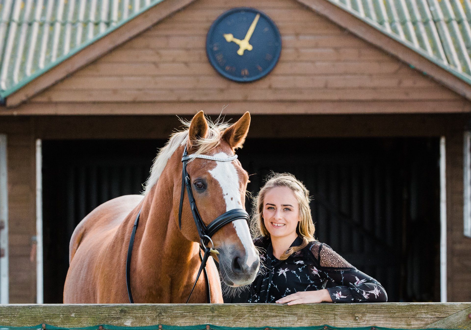 Woman and chestnut horse, posing together at stable, wooden building with clock in background.