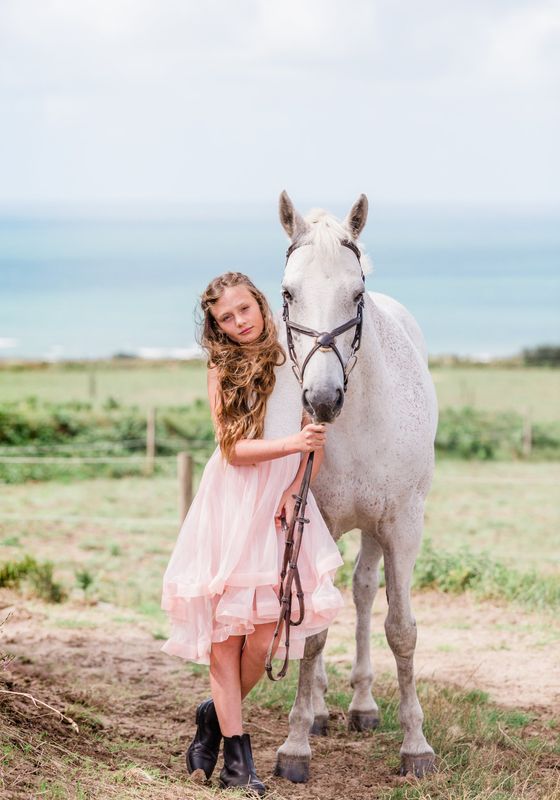 Girl in pink dress stands next to a white speckled horse outdoors near the ocean.
