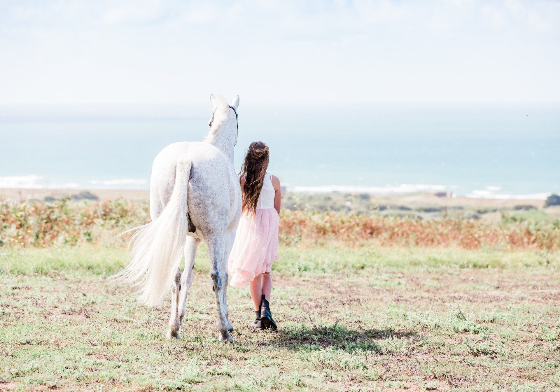 Girl in pink skirt walks with white horse on a grassy field, ocean in the background.