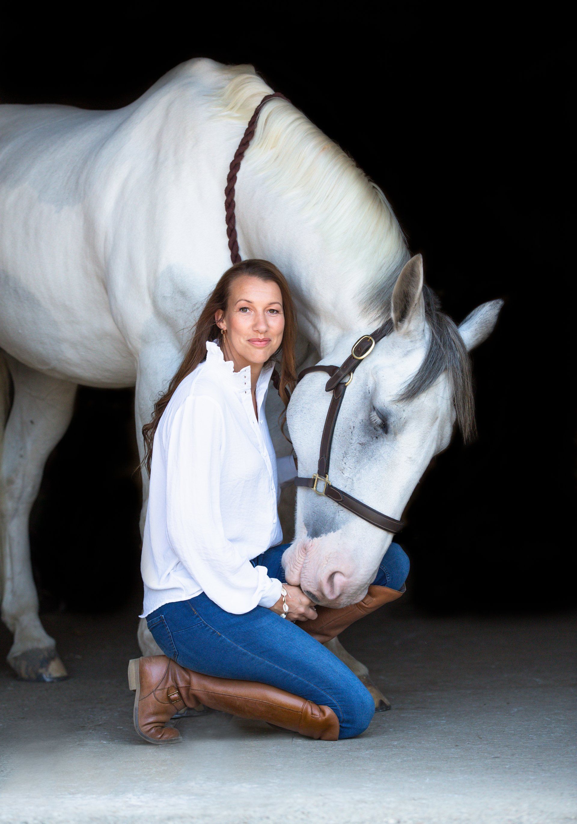 Woman kneeling next to a white horse in a stable, wearing a white shirt, jeans, and boots; both are looking at the camera.