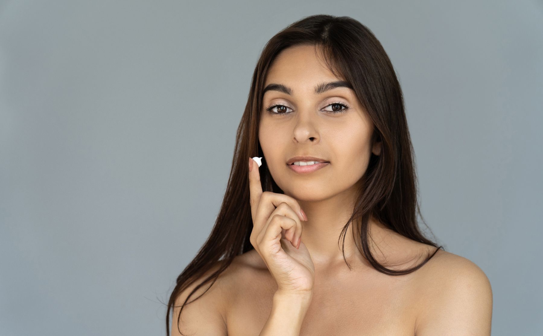 Woman with long dark hair posing against a gray background, touching her chin with one finger.