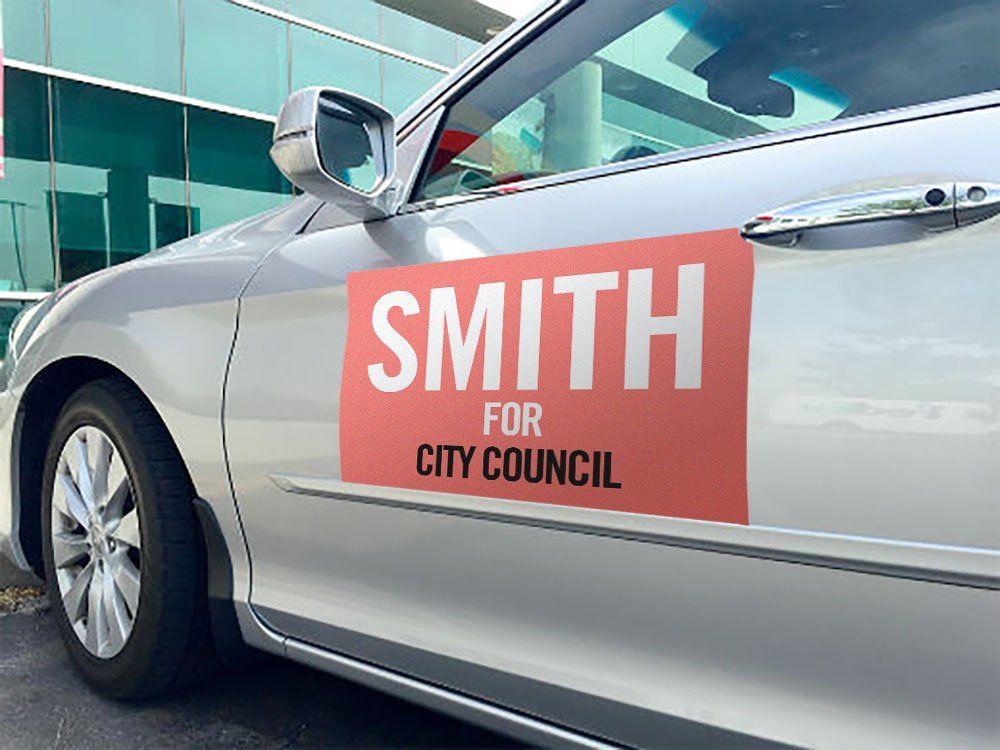 Silver car with red “SMITH FOR CITY COUNCIL” campaign sign on the door near a glass building