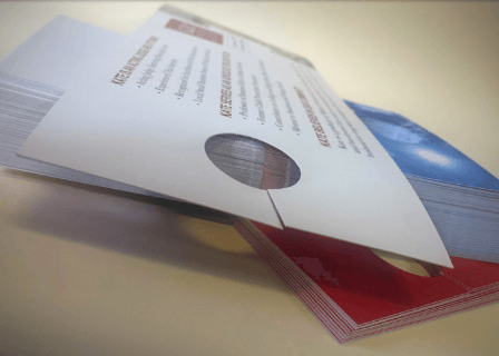 Stacked brochures and flyers on a table, with a white top sheet and red and blue papers beneath.