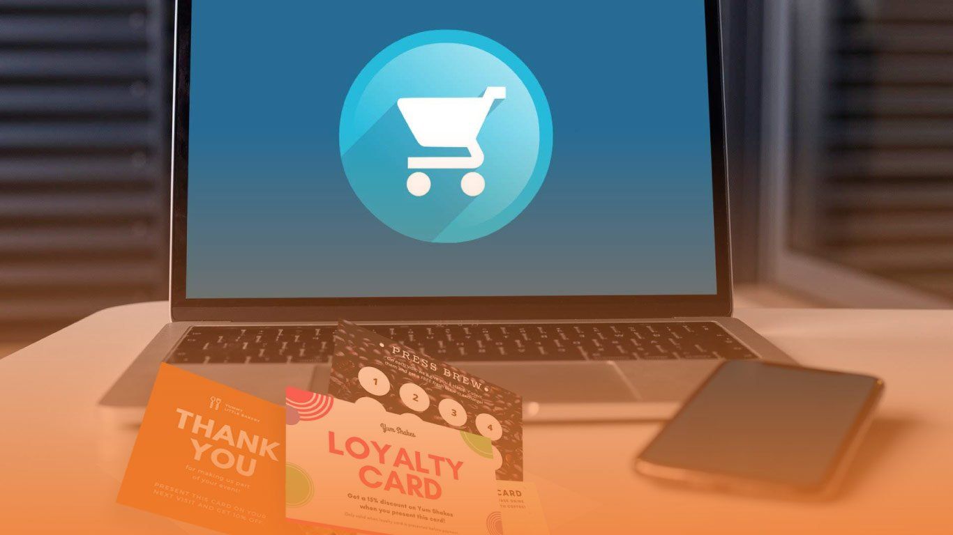 Laptop with shopping cart icon, surrounded by loyalty cards and a thank-you card on a desk.
