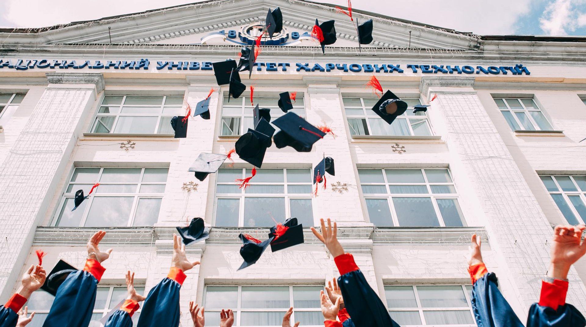 Graduates toss black caps in front of a white school building with blue lettering
