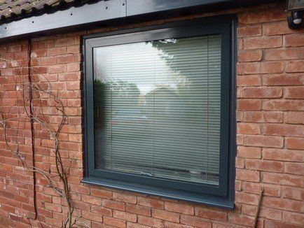 A dark gray square window frame with integrated horizontal blinds, set into a red brick exterior wall.