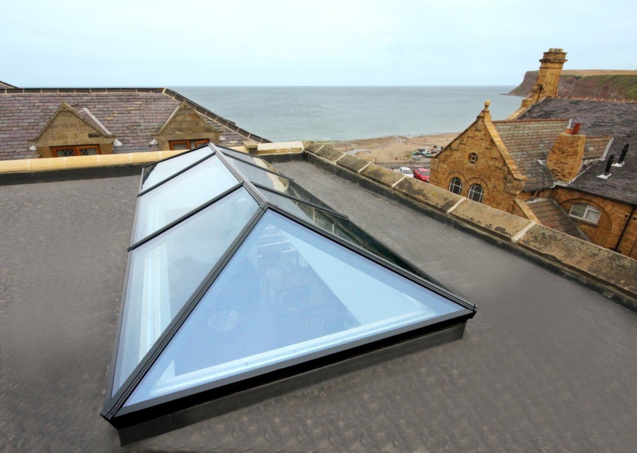 A glass lantern skylight sits on a flat, dark roof overlooking a coastal village and the sea.