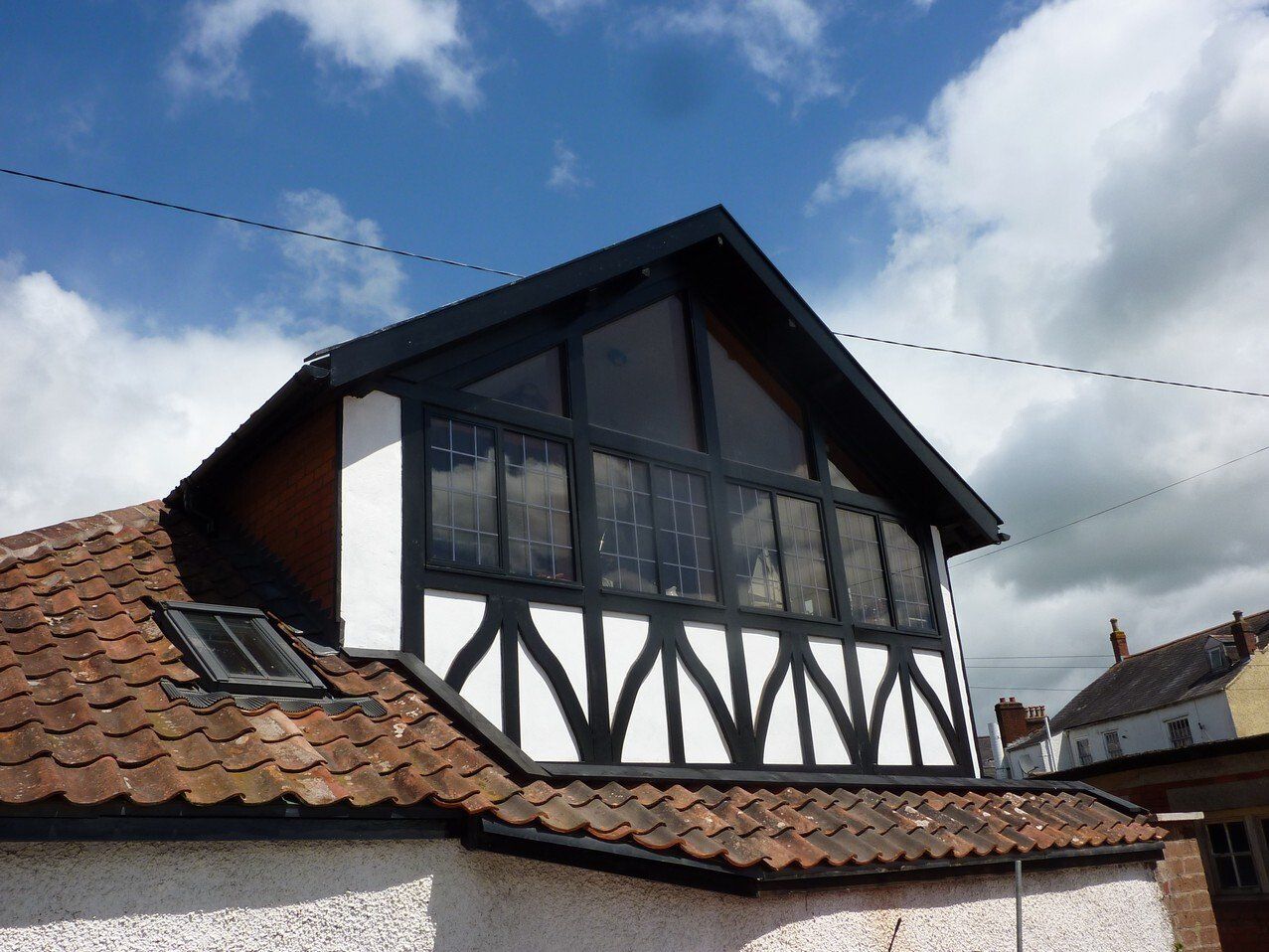 A gabled dormer addition with black timber framing and white infill, set against a red tiled roof and a blue cloudy sky.