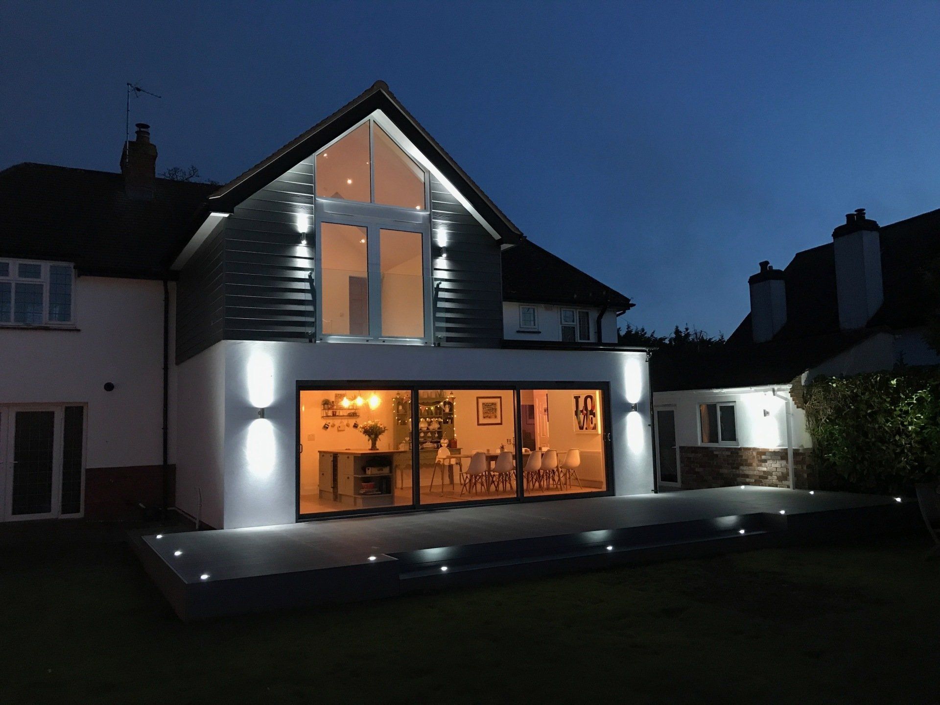 A modern house extension at dusk with lit floor-to-ceiling glass doors, a bright patio, and a gabled window above.