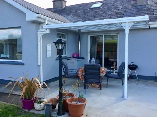 A grey house patio with a white pergola, outdoor table and chairs, potted plants, a lamp post, and a small grill.
