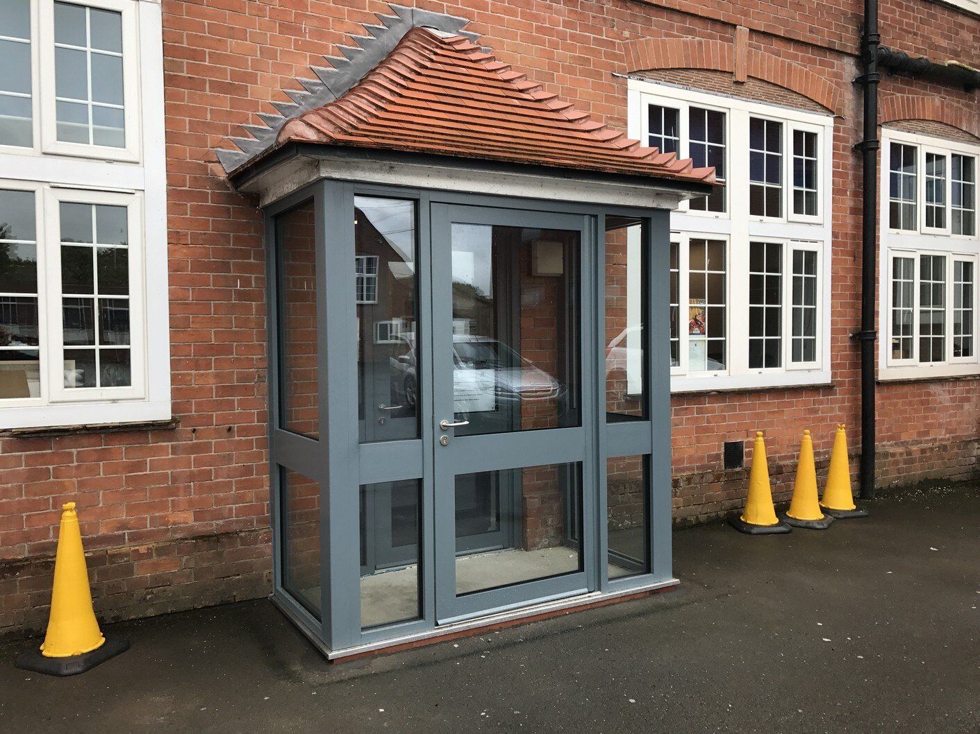 A grey, glass-walled entrance porch with a tiled roof attached to a brick building, with yellow traffic cones nearby.