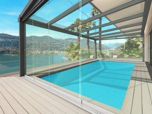 A rectangular swimming pool on a wooden deck under a glass-enclosed patio cover, overlooking a lake and mountains.
