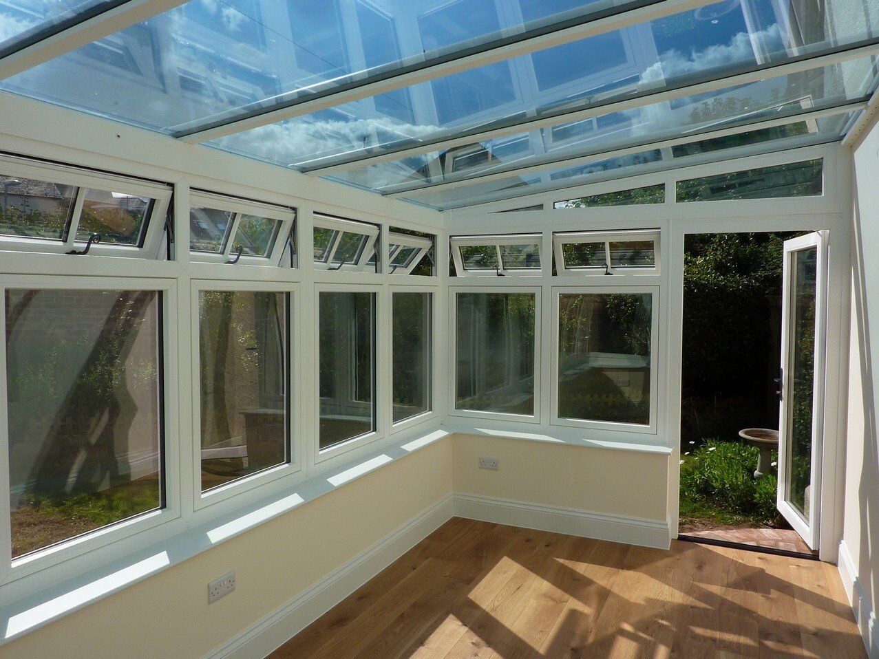 A bright, modern conservatory with a glass roof, white frames, light walls, and wood flooring, opening to a green garden.