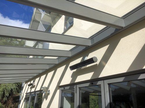 A glass-covered patio roof attached to a beige house, featuring a mounted outdoor heater on the wall above glass doors.