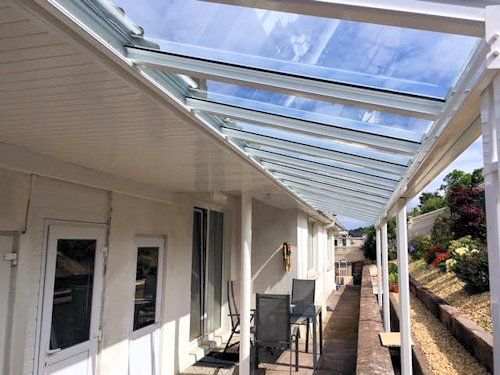 A covered patio featuring a white frame structure with a glass roof, adjacent to a house wall, overlooking a garden area.