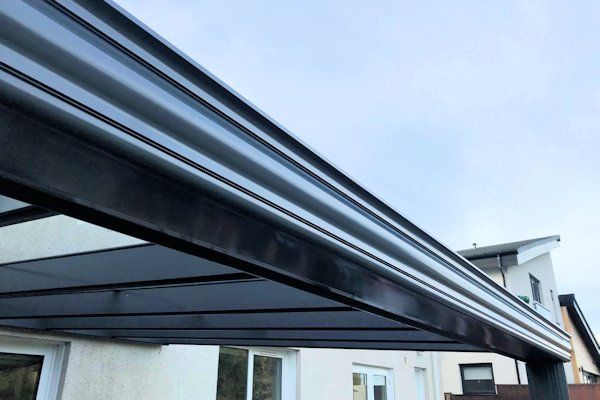 A low-angle view of a dark metal patio cover roof extension attached to the side of a light-colored house.