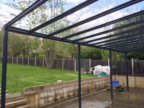 Black metal pergola with a transparent roof over a patio, overlooking a backyard with a lawn and fence.