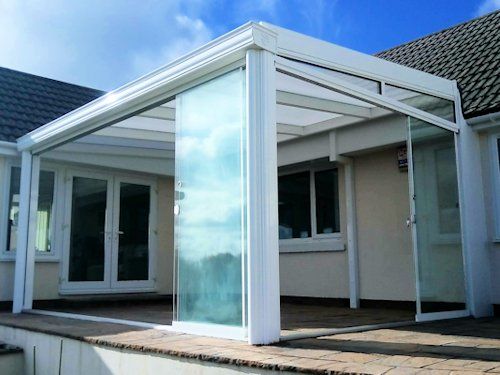 A white-framed glass patio enclosure attached to a house, featuring sliding glass doors over a paved patio area.