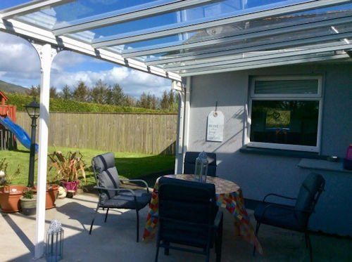 A white metal patio cover shelters a gray concrete area with a round table, three dark chairs, and potted plants.
