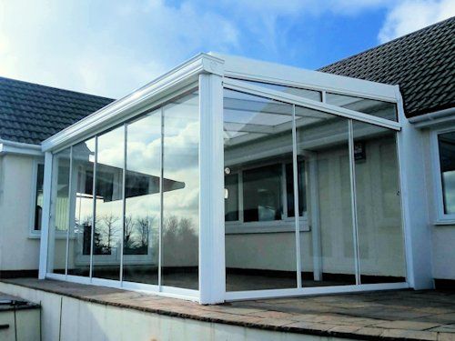 A white, glass-enclosed patio sunroom attached to the side of a house with a dark tiled roof.