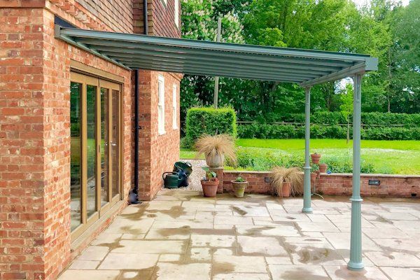 A patio with a green metal veranda roof attached to a brick house, overlooking a green garden with potted plants.